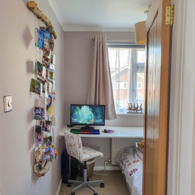 A small, tidy bedroom with a desk under a window and a computer setup. Woodland wall art and photos hang on the left wall, whilst a single bed with a patterned chair sits on the right. Sunlight filters through light curtains.