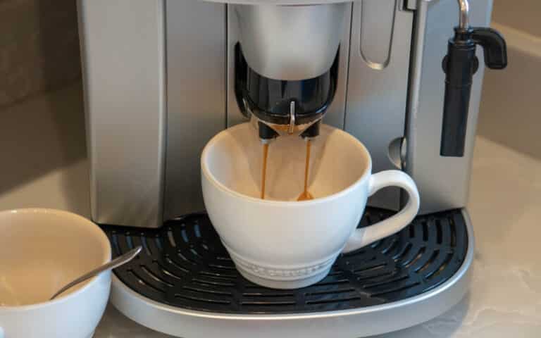 A white coffee cup sits under a coffee machine as espresso pours in, while a second cup with a spoon rests nearby. Perfect for sampling new blends from your Blue Coffee Box coffee subscription.