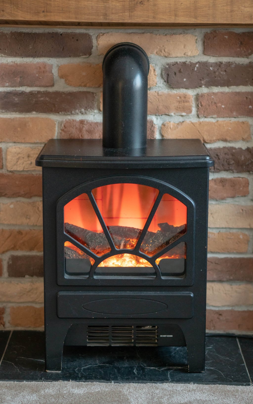 A black wood-burning stove with a glowing fire inside is set against a Kuci Design Brick Slip Fireplace. The stove features a glass front with decorative metal bars and a sleek black chimney pipe extending upwards.
