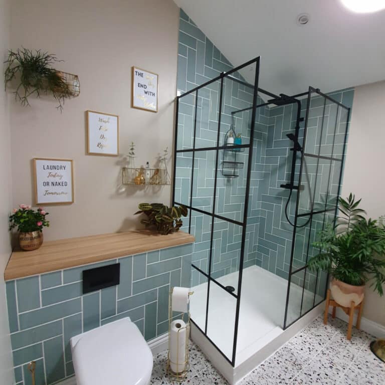Modern bathroom with green subway tiles in the shower, black-framed glass enclosure, wall art, and plants. A wooden shelf holds decor whilst a toilet roll holder stands nearby—perfect inspiration for your next Utility Room Tour.