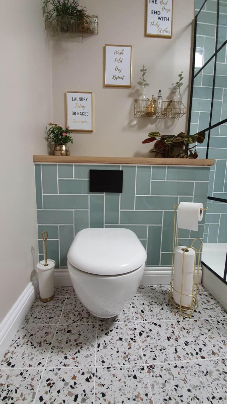 Modern bathroom with a floating white toilet, gold toilet roll holder, green tiles, terrazzo floor, wall shelves with decor, small plants, and framed quotes—offering utility room inspiration in every detail.
