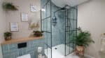 Modern bathroom featured in our House Tour, with light blue tiled shower, black-framed glass doors, and potted plants. Floating shelf holds decor and toilet roll. Motivational wall art hangs above the toilet for a light and airy vibe.