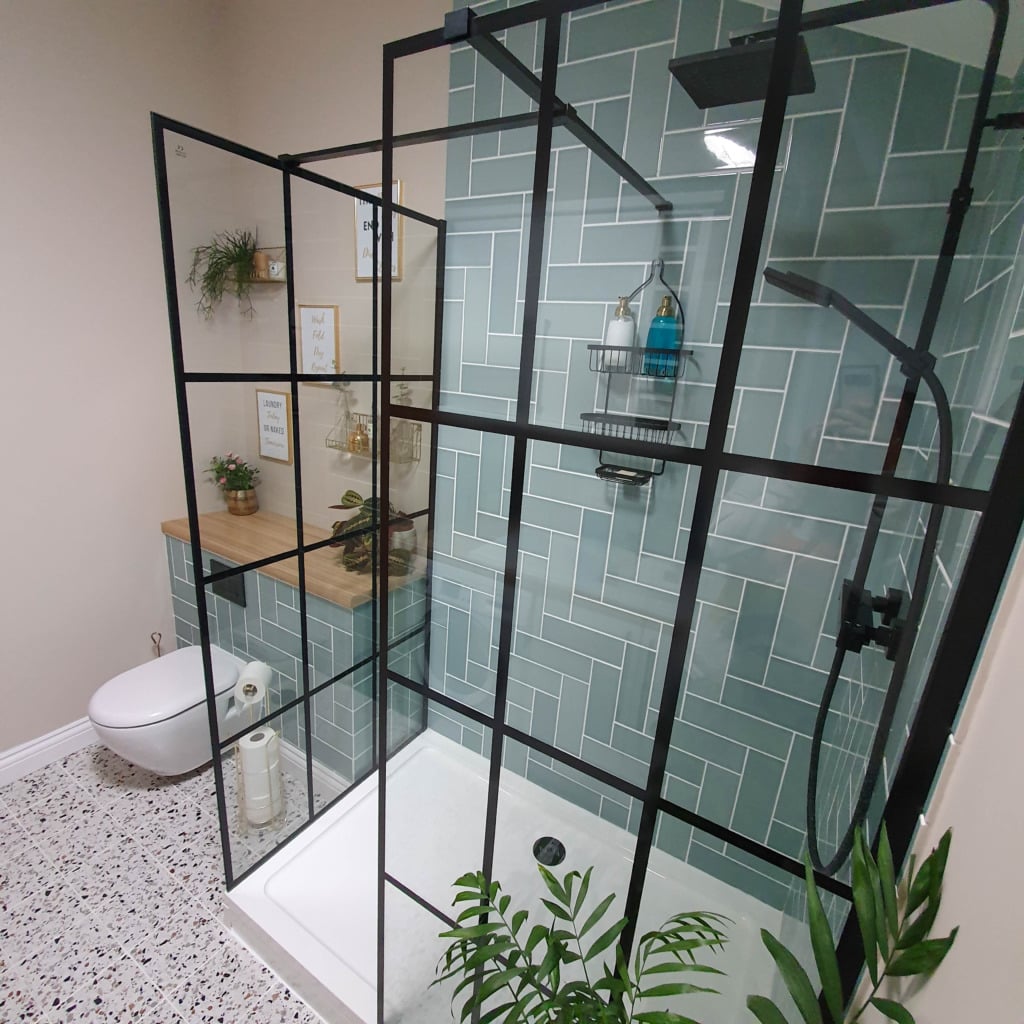 Modern bathroom with a glass shower enclosure, green herringbone tile wall, floating toilet, wooden countertop with potted plants, and terrazzo floor. Framed art and greenery add charm—perfect inspiration for Utility Room organisation.