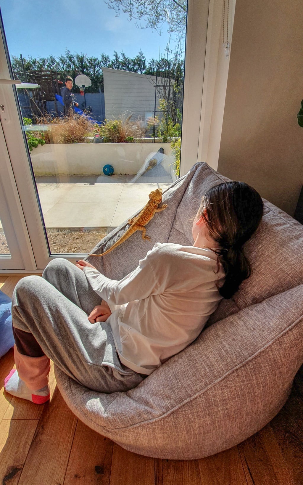 A girl relaxes on a light grey Beanbag Armchair indoors with a bearded dragon on her shoulder, sunlight streaming in. Outside, two people stand near a basketball hoop and a blue ball—just another cosy day from Beanbag Bazaar.