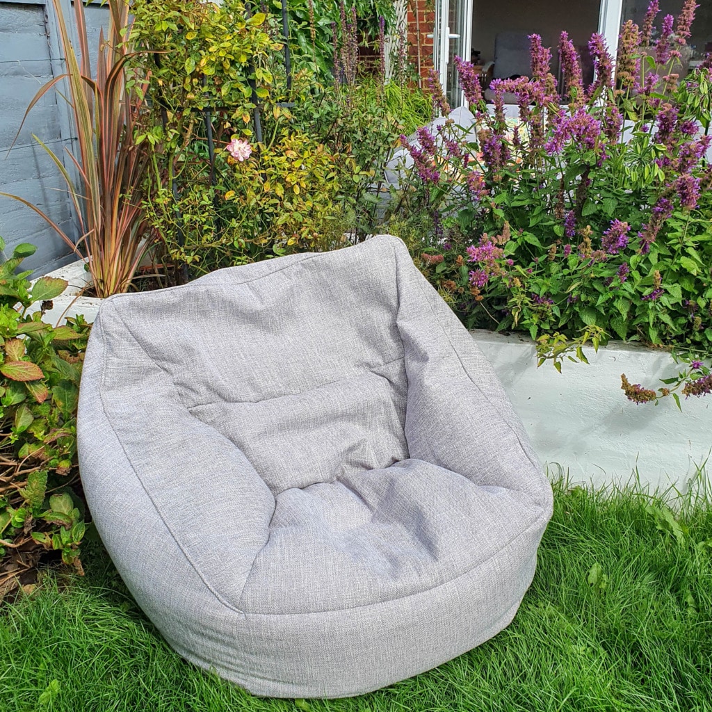 A light grey Beanbag Armchair from Beanbag Bazaar sits on green grass in a garden, surrounded by lush plants and purple flowers, with a patio and open doors visible in the background.