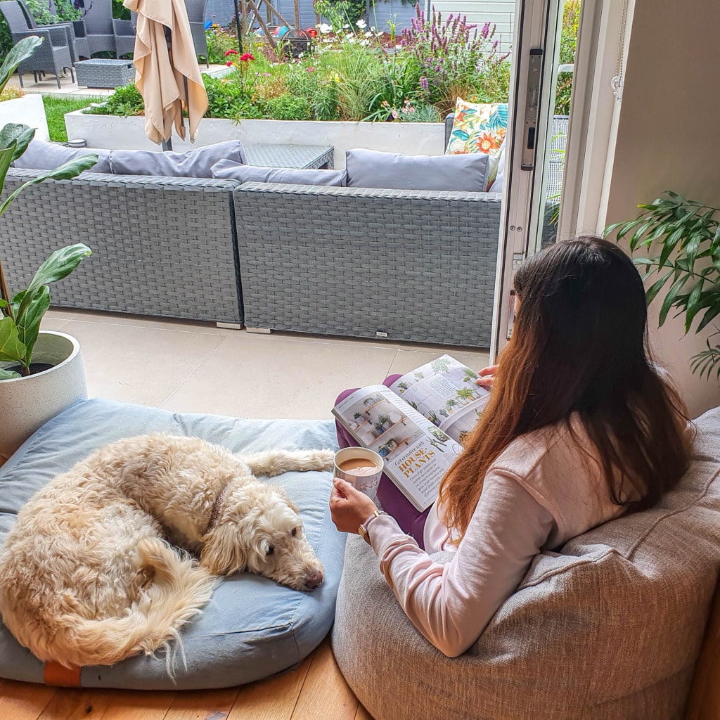 A woman sits indoors on a Beanbag Bazaar bean bag armchair, holding a mug and reading a magazine review. A fluffy dog lies on a cushion beside her, whilst a patio with grey furniture and a colourful garden is visible through open doors.