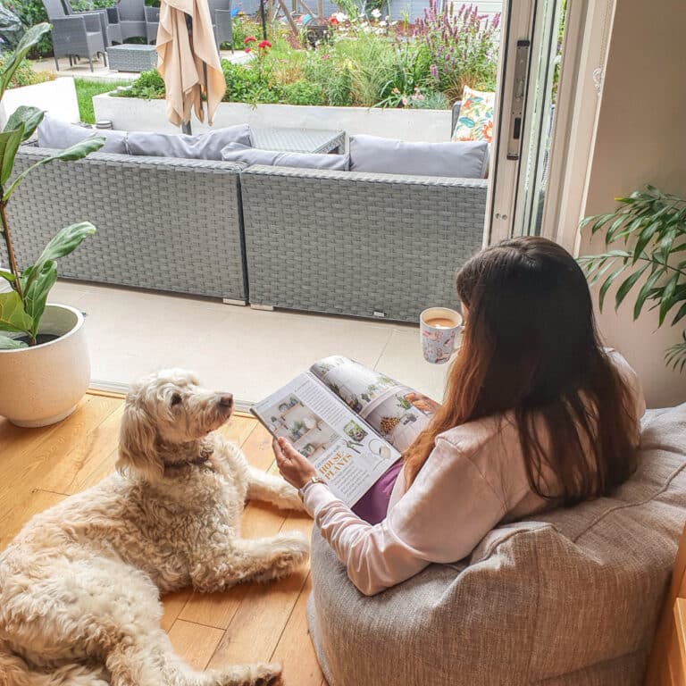 A woman relaxes on a Beanbag Armchair by an open door, reading a magazine and holding a mug. A fluffy dog lies beside her, with views of the patio and garden featuring outdoor furniture. Perfect for a cosy Beanbag Review moment.
