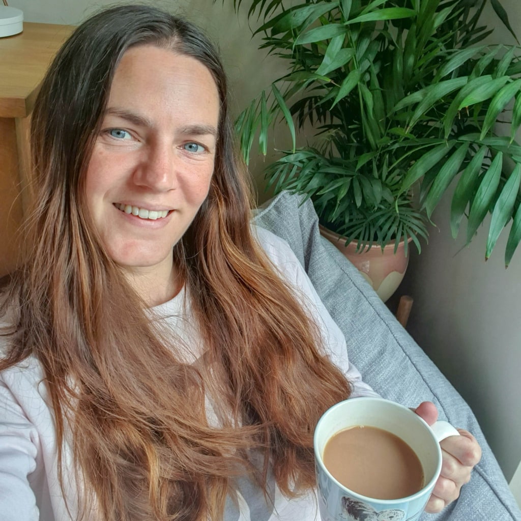 A woman with long brown hair sits on a grey Beanbag Armchair, smiling and holding a mug of tea or coffee. A leafy green plant in a pot rests on a stand beside her, creating the perfect spot to relax and unwind—worthy of any Beanbag Bazaar review.