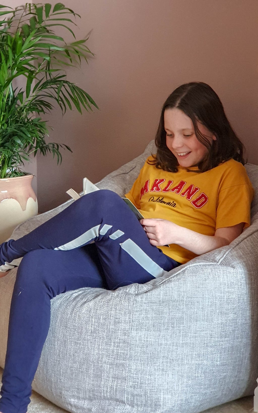 A smiling child with shoulder-length brown hair sits on a grey Beanbag Armchair from Beanbag Bazaar, wearing a yellow Oakland T-shirt and blue trousers, reading a book. A green potted plant is visible beside them.