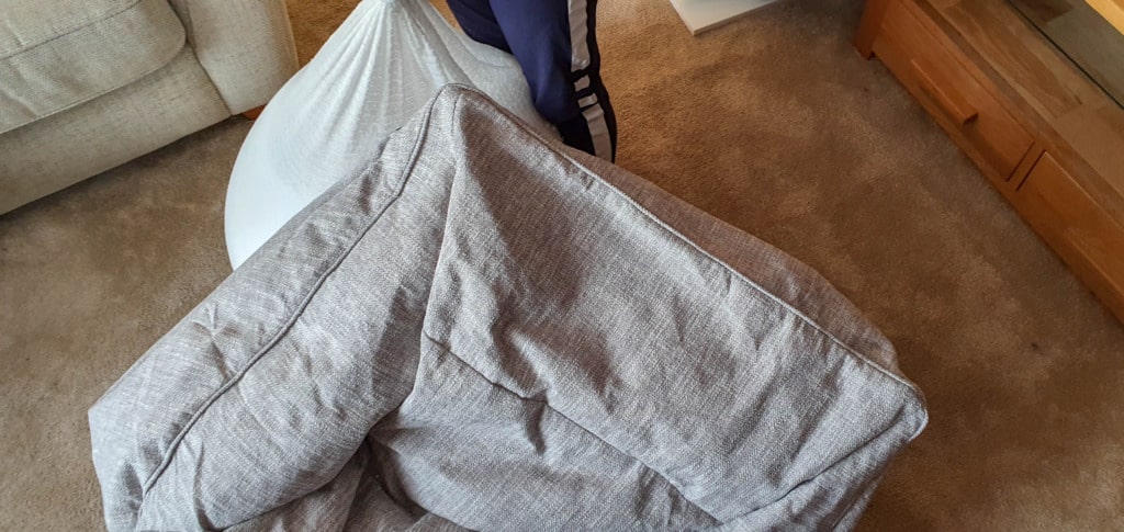 A Beanbag Bazaar bean bag chair is tipped over onto its side on a brown carpet, next to a light-coloured sofa and a wooden cupboard. Someone wearing blue trousers with white stripes is partially visible in this cosy living space.