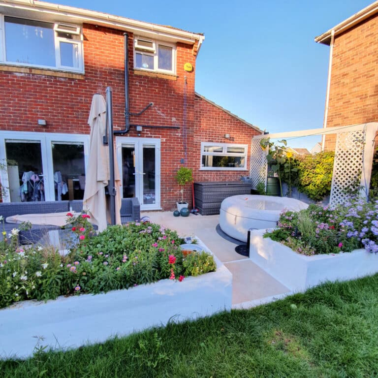 A back garden patio with outdoor furniture, a large parasol, a round hot tub, and raised flower beds filled with colourful blooms. The patio connects to a two-storey brick house, making My Garden the perfect August retreat on a sunny day.