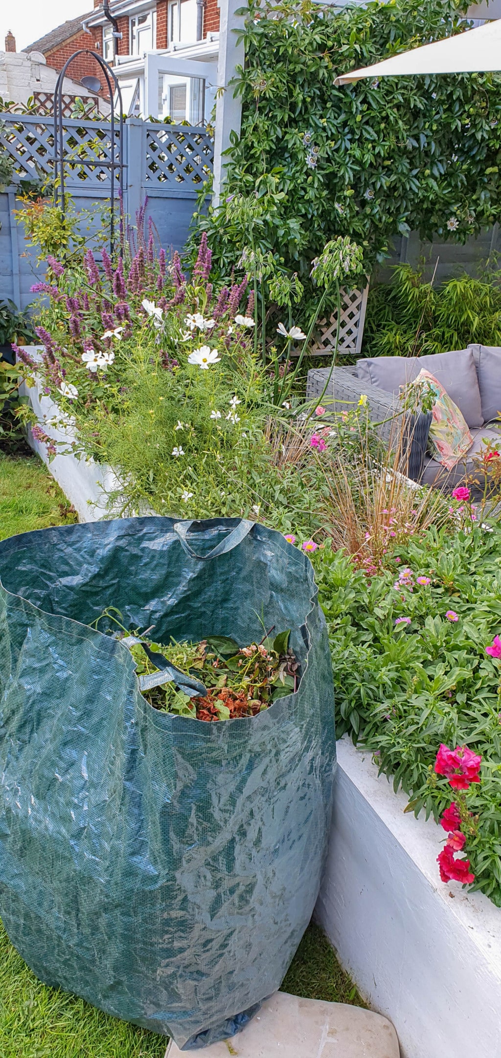 A large green garden waste bag filled with plant clippings stands on grass beside a raised flower bed in a neatly landscaped garden, with colourful flowers, lush greenery, and a seating area partially visible in the August sunshine.