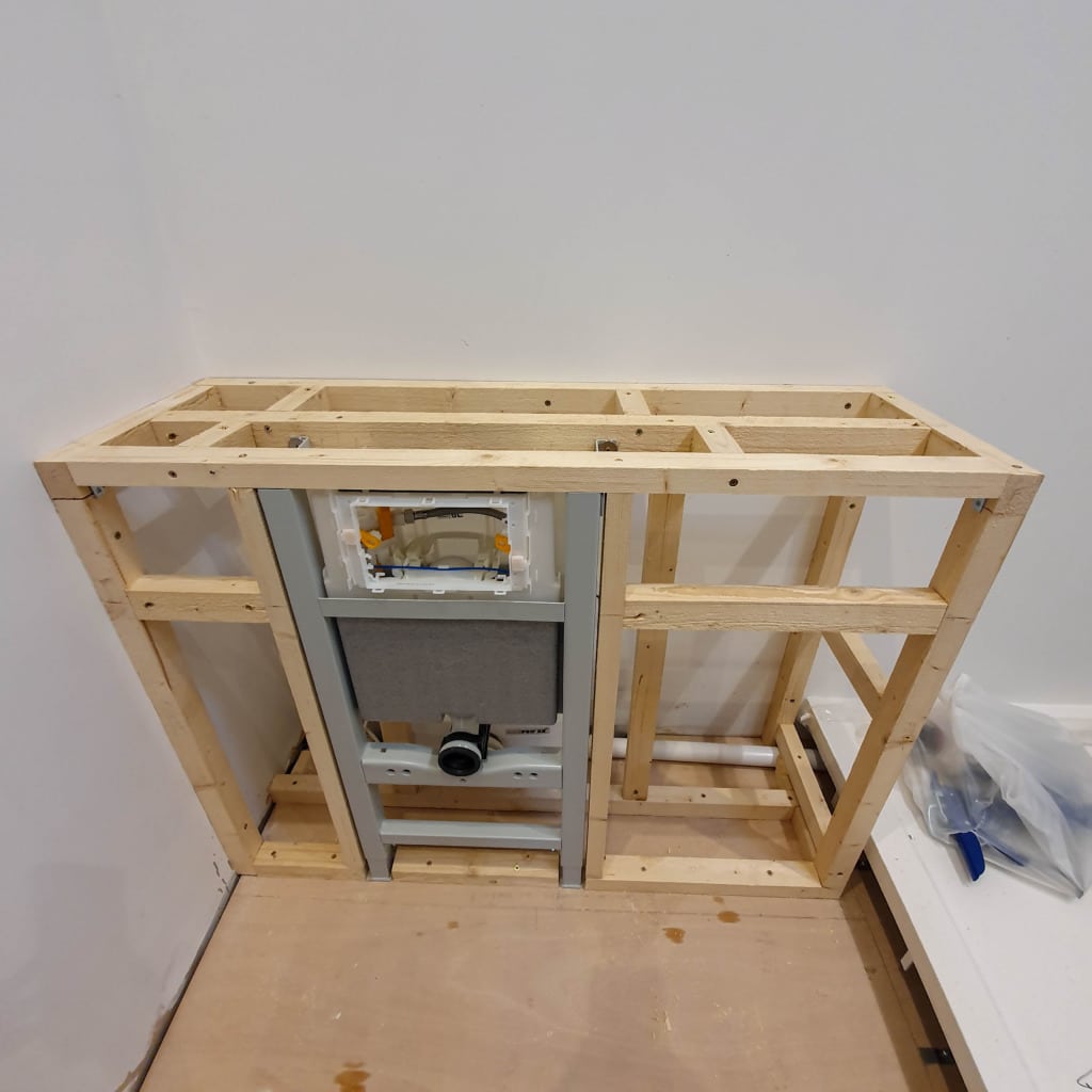 A wooden frame structure built against a white wall, enclosing a grey metal toilet cistern system—perfect for Utility Room Organisation—with tools and hardware in a bag on the floor to the right.
