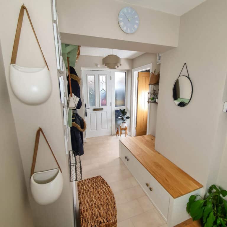 A tidy, light-filled neutral hallway with beige walls, a white front door, wooden bench with storage, round mirror, hanging planters, and a woven basket. Perfect for a Neutral Home Décor lover’s Hallway Tour—a blue clock and plant complete the entrance.