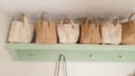 Five woven straw handbags are neatly lined up on a light green wooden shelf in a neutral hallway, each with short handles and a subtle herringbone or plaited pattern—perfect for a stylish hallway tour.