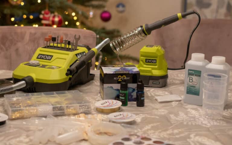 A table with resin craft supplies for Resin Christmas Crafts, featuring bottles of epoxy resin, pigments, moulds, a Ryobi soldering iron, and tools, set in a festive room with a decorated Christmas tree in the background.