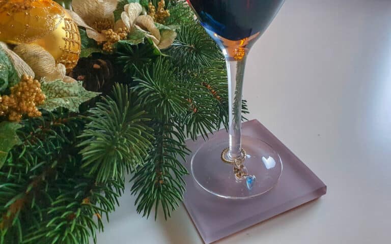 A wine glass with a charm on the base sits on a coaster near a festive holiday arrangement featuring pine branches, gold decorations, and a resin Christmas crafts ornament on a white surface.
