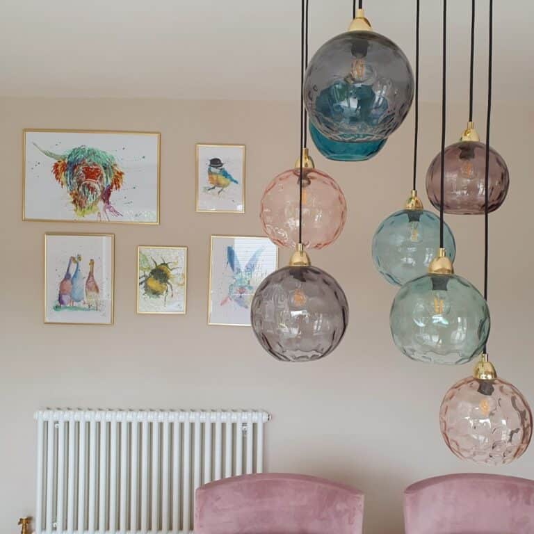 Colourful glass pendant lights hang in front of a beige wall featuring a stylish wall art installation with framed animal artwork. Two pink velvet chairs sit below, and a white radiator is visible on the left.