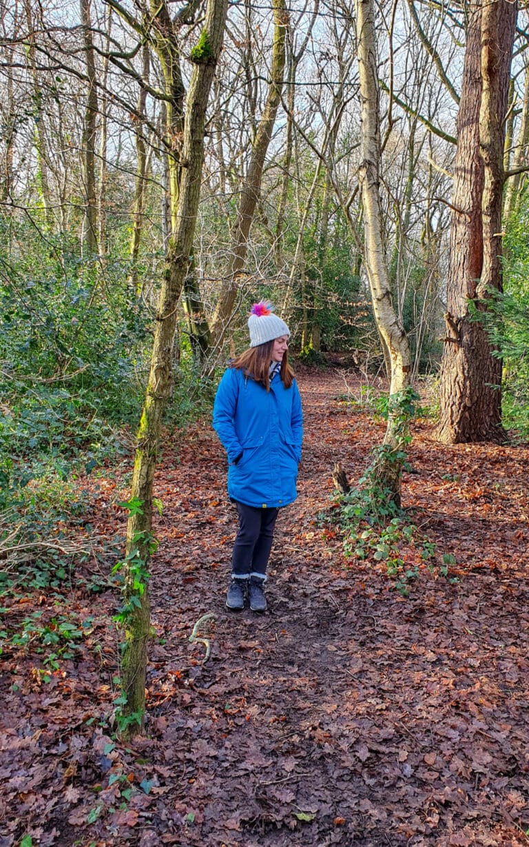 A person in a blue coat and grey hat stands on a leaf-covered path in a forest with bare trees, enjoying an outdoor activity on a sunny day—perfect for the 1000 Hours Outside Challenge.
