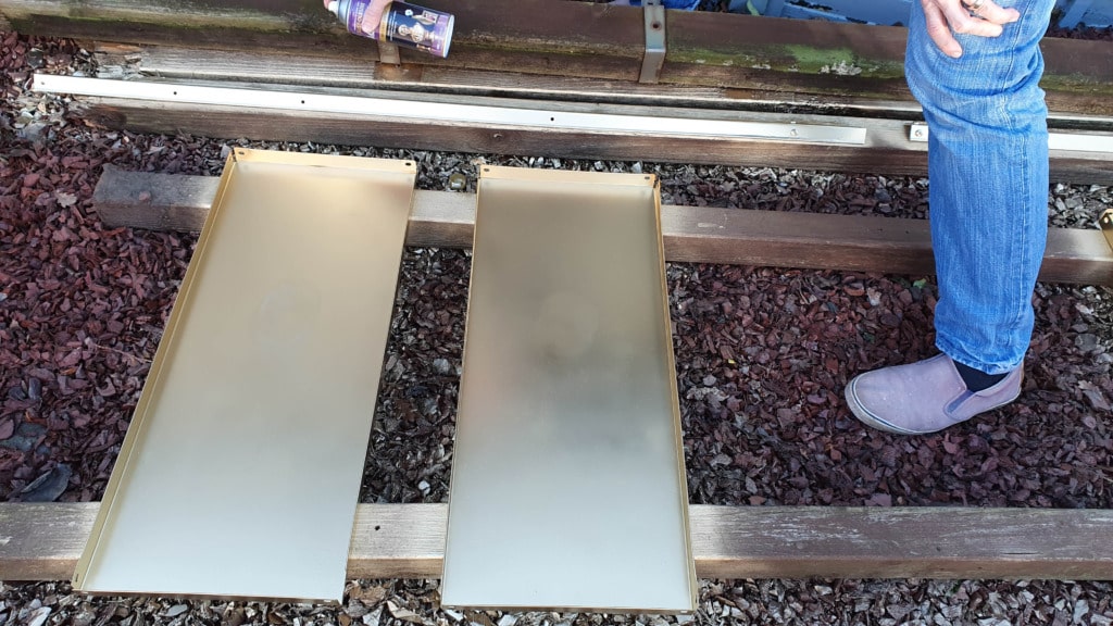 Two metallic panels lie side by side on wooden beams over gravel, the groundwork for a budget shelving unit. A person wearing blue jeans and light-coloured shoes stands to the right, partially visible.