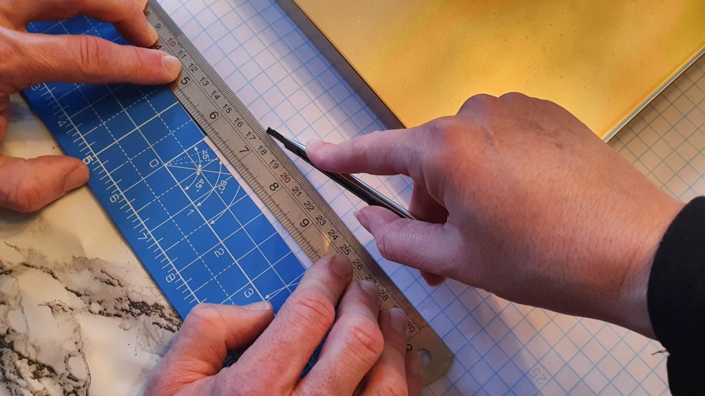 Two hands use a metal ruler and utility knife to cut blue grid-patterned paper on a larger sheet of graph paper, with a gold notebook nearby—perfect for planning budget shelving or your next DIY gold shelving project.