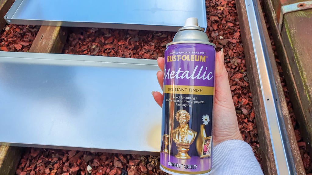 A hand holds a can of Rust-Oleum Metallic spray paint in front of metal trays on red gravel outdoors, perfect for transforming budget shelving or a DIY shelving unit with a brilliant, bright gold finish.
