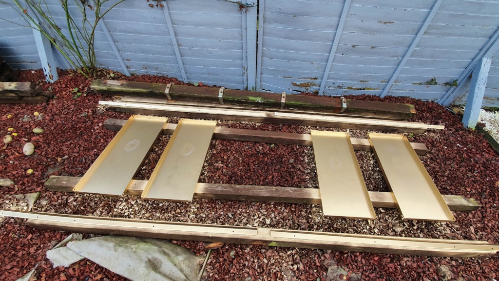 Four rectangular golden panels, resembling DIY gold shelving, rest on wooden beams laid out on a bed of red gravel in a garden, with a blue wooden fence in the background.