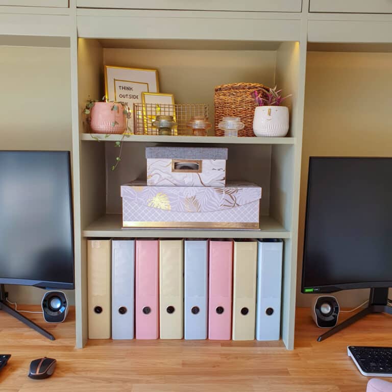A tidy desk shelf with built-in storage holds two monitors, colourful file folders, decorative boxes, potted plants, candles, and framed art. The soft pastel palette and organised setup create a cosy feel.