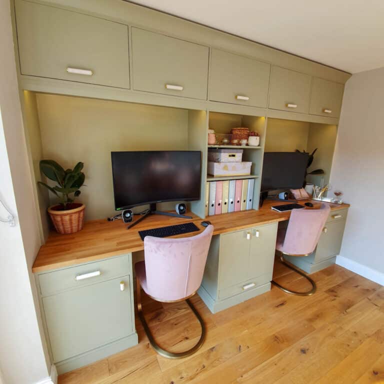 A tidy home office with two DIY built-in desk workstations, each featuring a monitor, keyboard, pink velvet chair, and potted plant, set against built-in green cupboards with shelves holding storage boxes and folders.