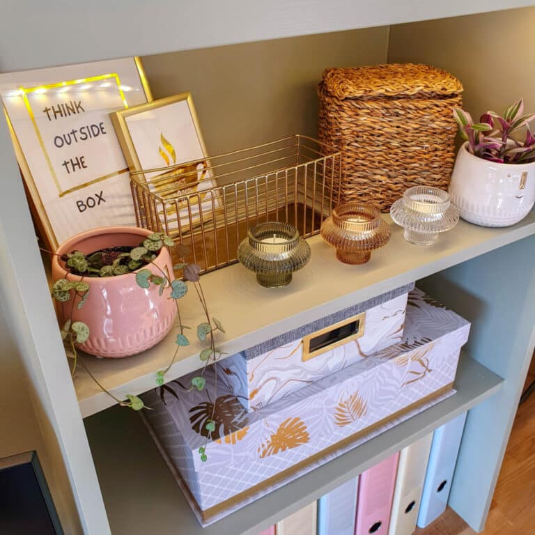 A neatly organised DIY desk shelf with decorative candles, a pink plant pot, a framed “Think Outside the Box” sign, a woven basket, a gold wire tray, a potted plant, and patterned storage boxes and folders.