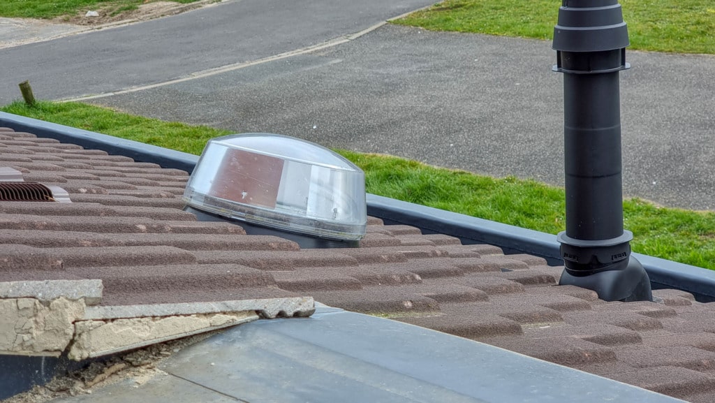 A round, clear Solatube dome skylight and a black ventilation pipe installed on a brown tiled roof—an ideal way to light a room with no windows. Grassy areas and a paved pavement are visible in the background.