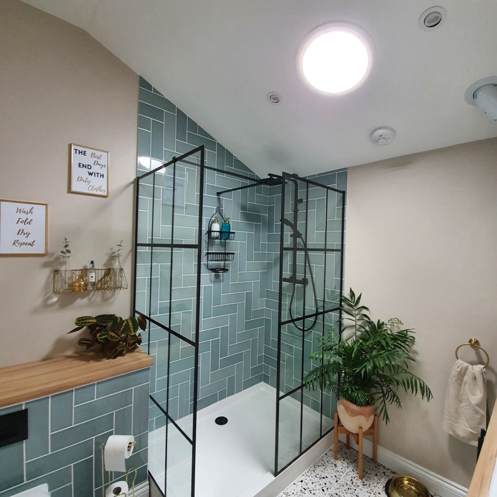 A modern bathroom with no windows features a glass shower enclosure, blue herringbone tiles, beige walls, a potted plant, and wall decor. A Solatube Sun Tunnel helps light a room filled with towel ring, toilet roll, and toiletries.