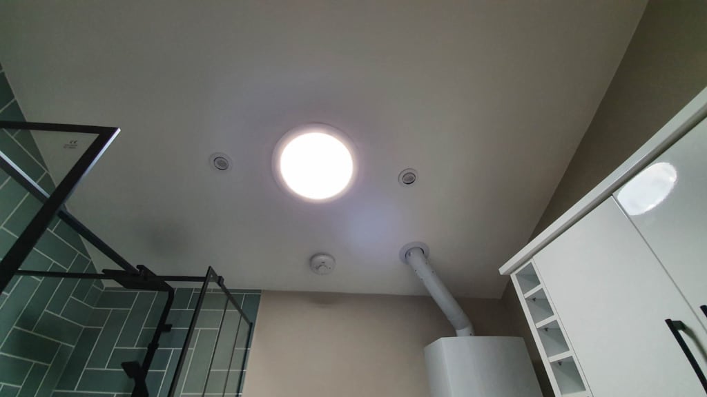 View of a bathroom ceiling with a round Solatube ceiling light to naturally light a room, ventilation ducts, a white water heater, green-tiled shower area, and white cupboards on the right side.