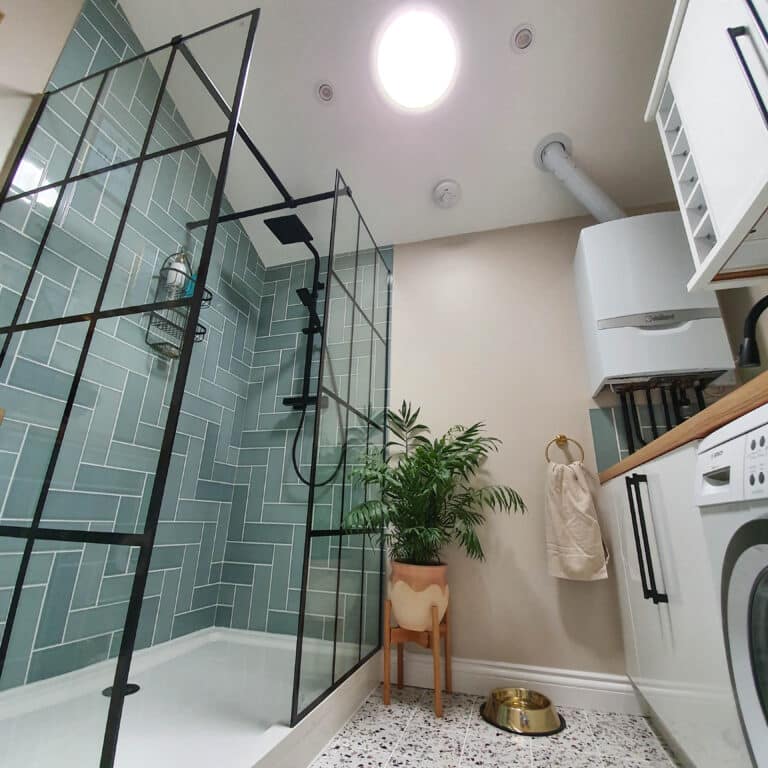 Modern bathroom with a green tiled shower, black-framed glass door, potted plant, white walls, boiler, towel on a gold ring, pet bowl on terrazzo floor, and a kitchenette with washing machine—naturally lit by a Solatube sun tunnel.