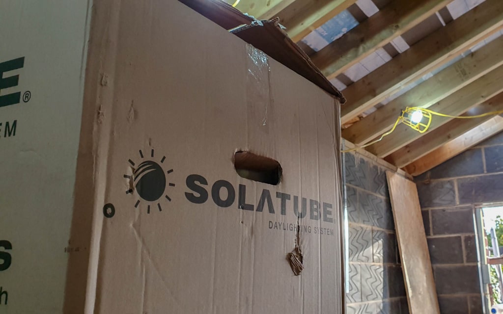 A large Solatube Daylighting System box stands upright inside a room under construction, with exposed wooden beams, breeze block walls, and a hanging work light—showing how to light a room with no windows using Sun Tunnel technology.