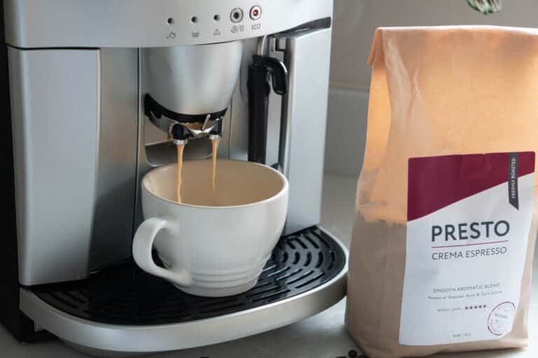 A white coffee cup sits under an espresso machine with coffee pouring into it. Next to the machine is a bag labelled PRESTO CREMA ESPRESSO, showcasing the rich flavour of Presto Coffee’s Crema Espresso Beans on a kitchen counter.