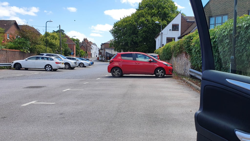 A red car is parked awkwardly across several empty spaces near a kerb in Bishops Waltham, with other cars properly parked nearby. The photo, taken from inside another car with its door frame visible, shows trees and houses in the background.