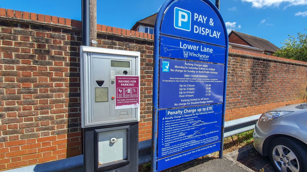 A parking meter and a blue Pay & Display sign in a Bishops Waltham car park next to a brick wall. The sign lists parking charges, hours, and penalties—perfect for anyone starting a Circular Walk nearby. Part of a parked silver car is visible on the right.