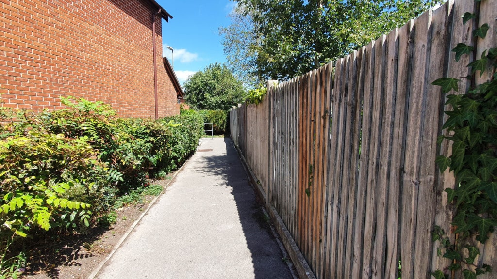 A narrow paved path, part of the Bishops Waltham Walk, runs between a brick building with green bushes and a tall wooden fence covered with ivy, leading towards trees in the distance under a blue sky.