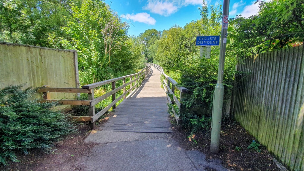 A wooden footbridge surrounded by greenery leads into a sunlit path on the Bishops Waltham Circular Walk. A blue sign on a lamp post reads “CYCLISTS DISMOUNT.” The sky is clear with a few clouds and fences border both sides of the path.