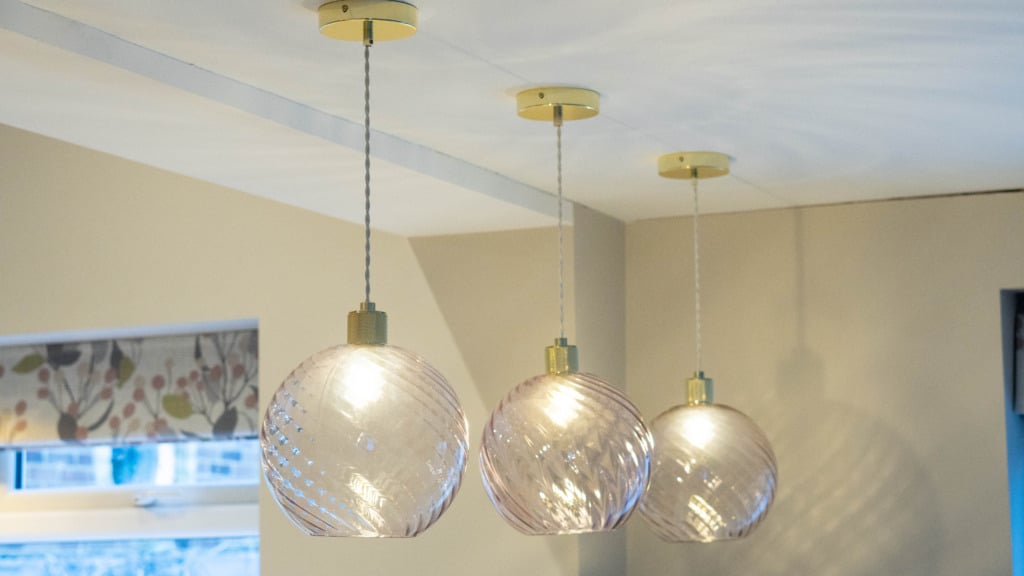 Three Creative Cables Lights with spiral glass pendants hang in a row from the ceiling, each illuminated, in a modern room with cream-coloured walls and patterned curtains—a perfect example of bespoke lighting.