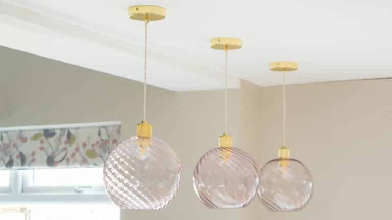 Three glass pendant lights from Creative Cables with gold fittings hang in a row. The glass shades feature a subtle spiral pattern, and a window with a floral patterned curtain is visible in the background.