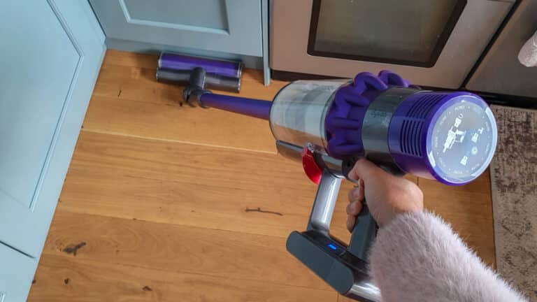 A person holding a cordless Dyson V10 vacuum cleaner is about to clean a wooden floor in a kitchen near blue cupboards and a stainless steel cooker.