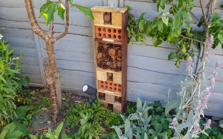 A homemade bug house crafted from wood, bricks, and pine cones stands upright in a garden by a grey wooden fence, surrounded by green plants and flowers—a perfect example of how to make an insect hotel using a pallet.