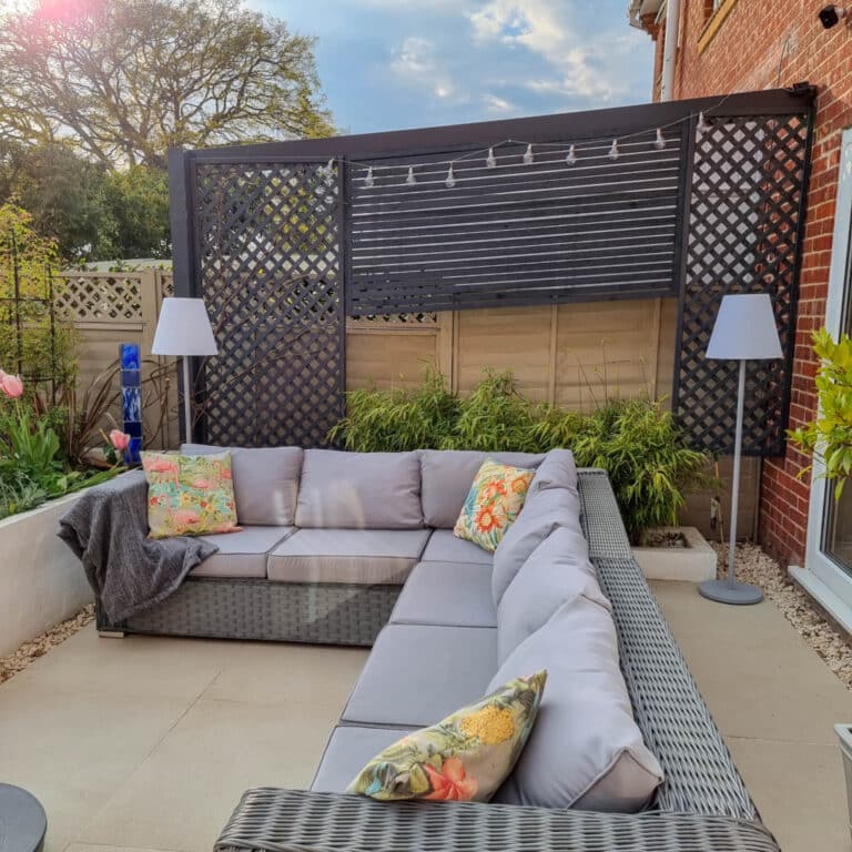 Large outdoor patio with an L-shaped grey wicker sofa, colourful floral cushions, a grey throw, tall garden lighting, black trellis privacy screen, green plants, and a brick wall on one side. Tulips and an evening garden are visible on the left.