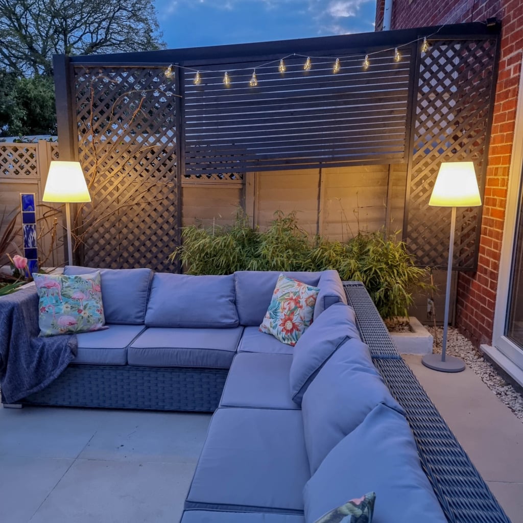 A cosy outdoor patio with a U-shaped wicker sofa, colourful cushions, two floor lamps, garden lighting, string lights, and green plants against a wooden lattice fence creates a warm, inviting evening garden atmosphere.