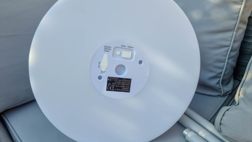A close-up of the bottom of a round white device, showing a label with technical information, a power port, and two Ethernet ports, resting on a light grey outdoor cushion in an evening garden with subtle outdoor lighting.