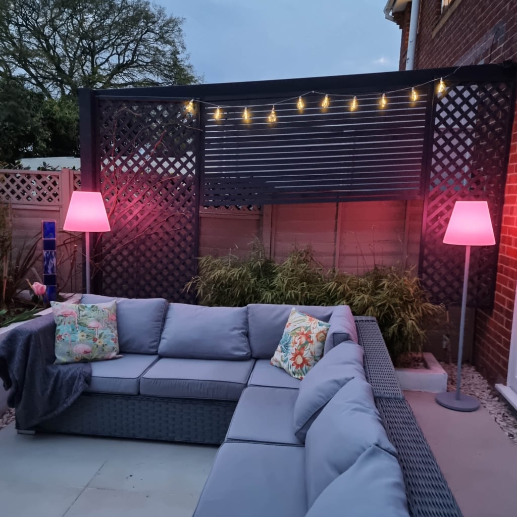 A cosy outdoor patio with a grey sectional sofa, colourful floral cushions, a blanket, and two pink-lit floor lamps. Fairy lights provide warm evening illumination above, while plants line the back fence under a trellis panel.