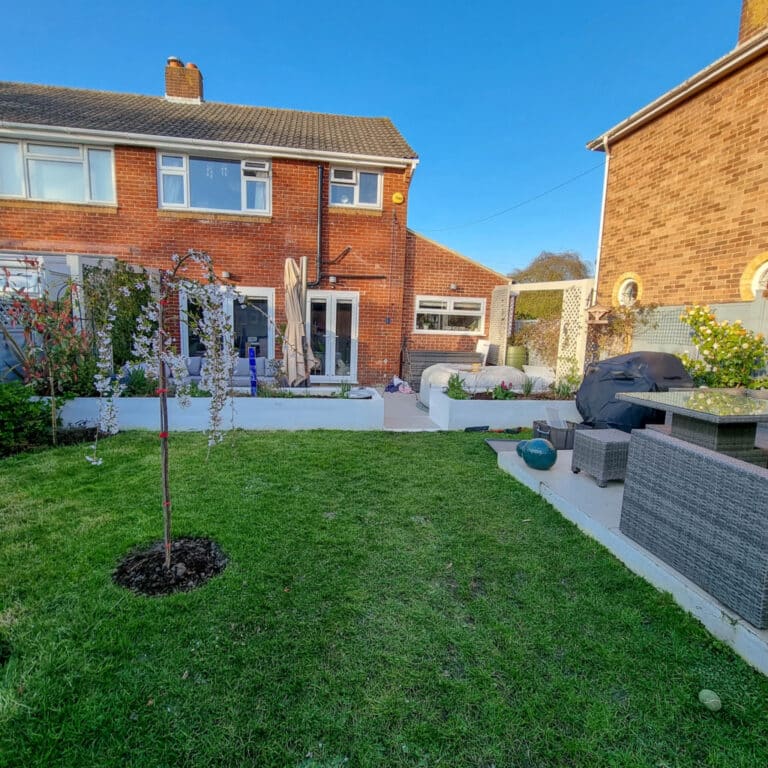 A neatly kept garden with green grass, a small tree, outdoor seating, a barbecue, and vibrant plants surrounds a brick semi-detached house with large windows—perfect for April gardening under a clear blue sky.