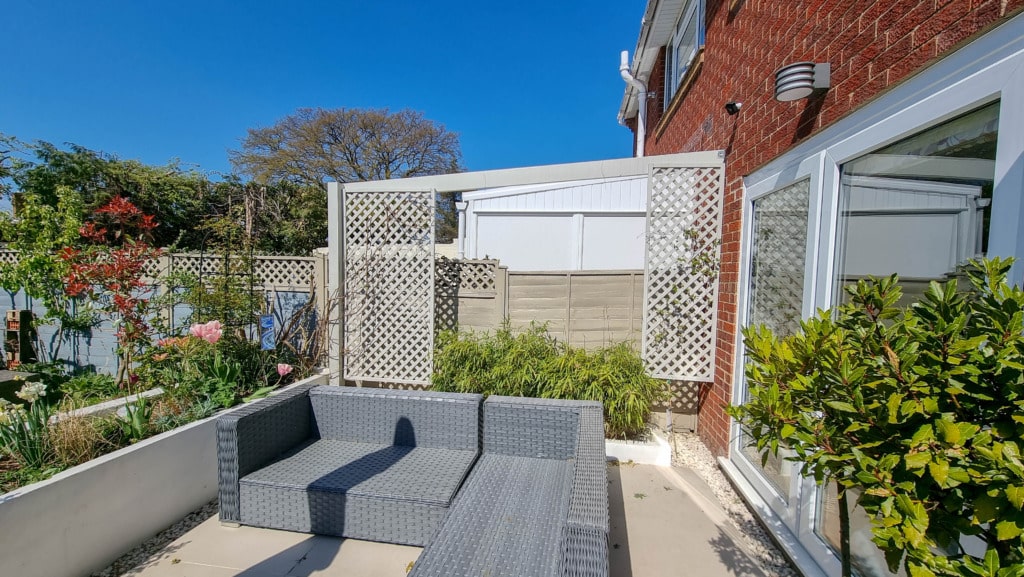 A modern outdoor patio with grey wicker seating, a red brick wall, large windows, green plants, and a blue sky on a sunny April day. There is a white lattice privacy screen and a garden bed perfect for gardening along the side.
